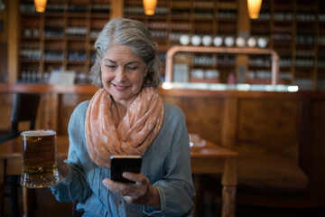 Senior woman wearing polka-dot scarf holding smartphone and glass beer mug at wooden table in pub