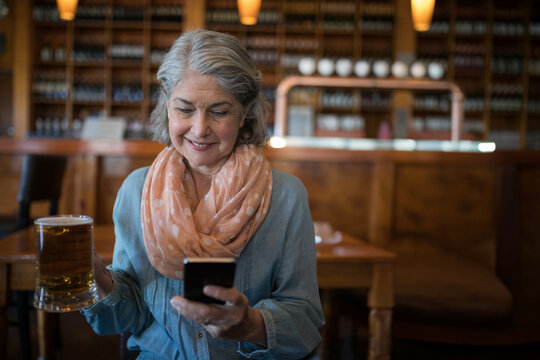 Senior woman wearing polka-dot scarf holding smartphone and glass beer mug at wooden table in pub - Powered by Adobe