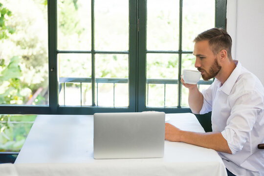 Man sipping coffee while working on silver laptop at dining table by window, copy space