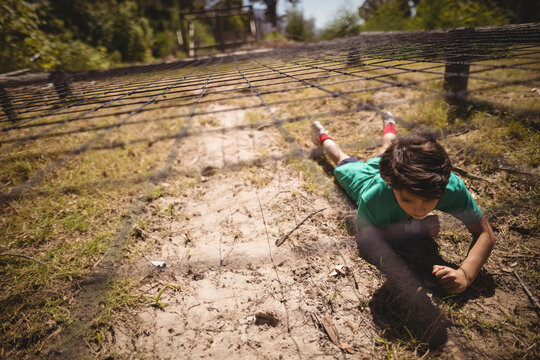 Boy is crawling under nylon obstacle net on sandy path casting grid shadows in wooded park