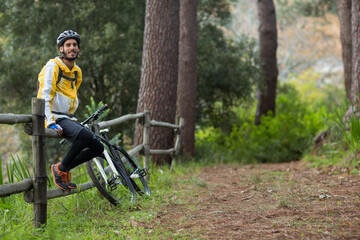 Man sitting on split-rail fence beside mountain bike on trail wearing helmet, daypack, copy space © wavebreak3