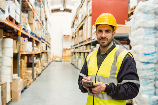 Male warehouse worker wearing vest scanning pallets on racks with scanner in aisle, copy space