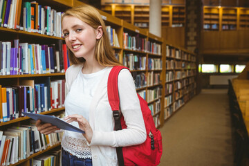 Teenage girl holding tablet and wearing red backpack in library aisle by bookshelves, copy space