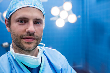 Surgeon wearing blue cap and scrubs with mask resting at chin under surgical lights