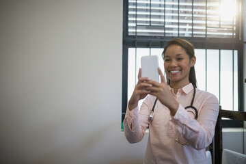 Mid adult professional holding smartphone with stethoscope by window blinds in office, copy space