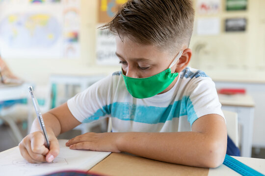 School-aged boy writing in notebook on classroom desk wearing green mask with pencil and ruler - Powered by Adobe