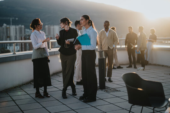 A group of diverse business people engaged in a brainstorming session on a rooftop at sunset. Multiracial colleagues collaborate, showcasing teamwork and creative ideas against a scenic city backdrop. - Powered by Adobe