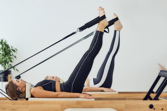 Two young women lying down on pilates reformer machine bed with legs up and feet in straps, working out and training at gym studio. Fitness, wellness concept 