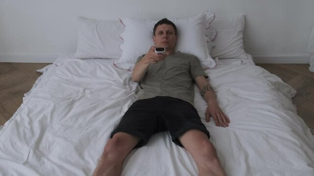 A young man lies on a bed holding a TV remote, switching channels in boredom. He spends time alone, feeling depressed and unmotivated.