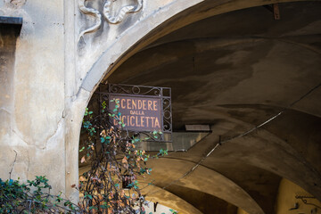 Reggio Emilia, Italy, 01.04.25. Rusty Italian sign in arched portico says dismount bicycle, surrounded by climbing ivy