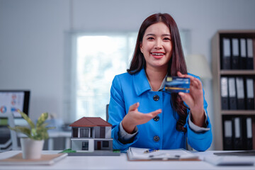 Smiling real estate agent showing credit card while sitting at desk with house model and contract,...