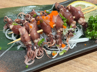 Hotaru ika sashimi skewer are elegantly presented on a black plate, accompanied by fresh vegetable and vibrant garnish, showcasing a culinary delight.