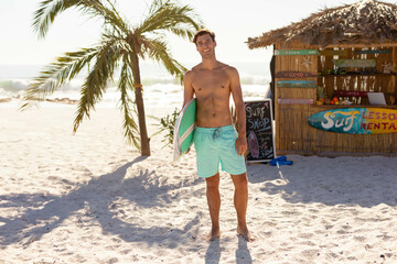 Shirtless man standing barefoot on beach holding surfboard beside surf rental hut with chalk sign