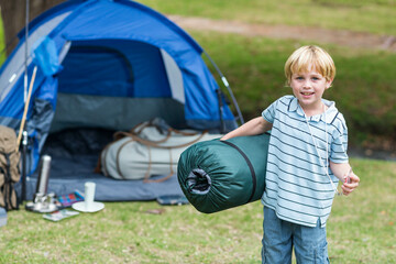 Boy carrying rolled sleeping bag while standing on grassy campsite by blue dome tent, copy space