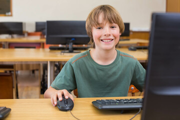Male child student smiling while using desktop computer and wired mouse at school computer lab desk