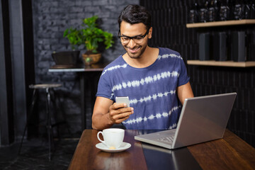 man is sitting at wooden table in café interior, holding smartphone beside laptop and coffee cup
