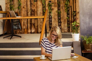 Female in her twenties typing on laptop at café table beside coffee cup pastry and smartphone