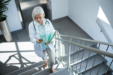 Senior female doctor climbing hospital stairs, holding smartphone and folder with stethoscope