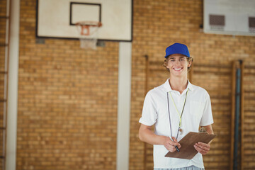 White teenage boy refereeing in gym holding clipboard, whistle on lanyard by hoop, copy space