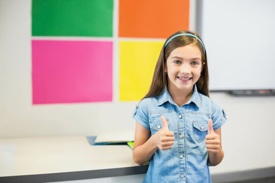 Girl student standing at school desk in bright classroom, holding blue notebook, giving thumbs up