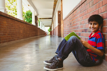 Hispanic boy reading green book while sitting on school corridor floor by brick wall, copy space