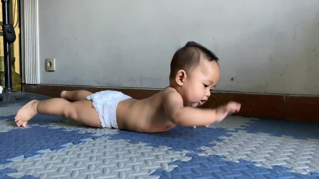 Infant boy, wearing only a white diaper, is in a tummy time position on a blue and gray interlocking foam mat in a house.