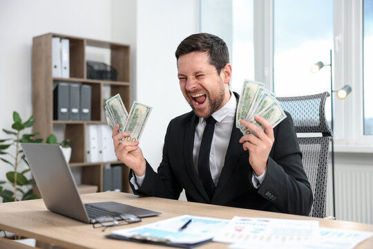 Happy greedy banker with dollar banknotes at table in office