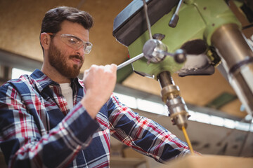 Bearded woodworker in safety glasses gripping drill press lever positioning plank in workshop