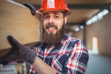 Worker carrying wooden beam on shoulder inside unfinished building, with red hard hat and gloves