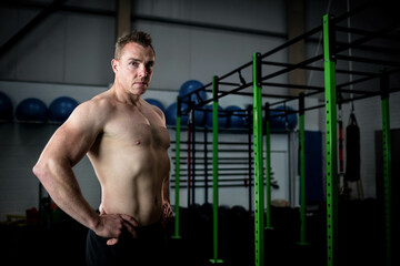 Shirtless muscular man standing with hands on hips in gym near green pull-up rig, copy space