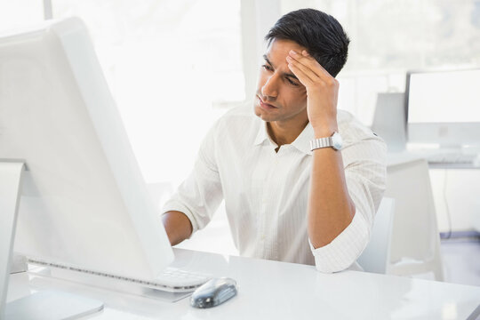 Indian man wearing collared shirt pressing hand to forehead at white desk facing computer monitor