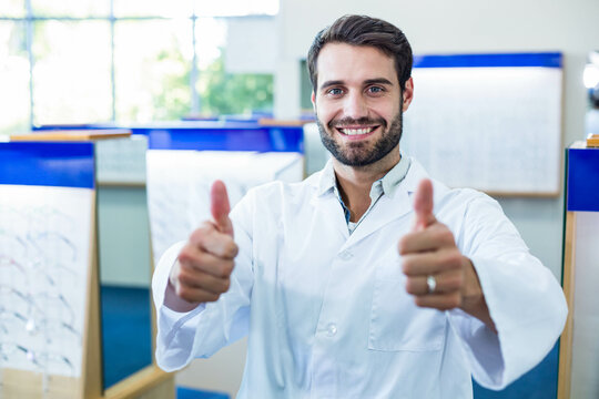 Man standing in optical shop wearing white lab coat, giving thumbs up at eyeglass display racks