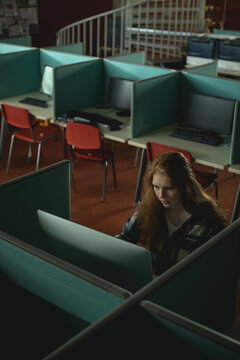 Female student typing on keyboard at computer lab cubicle by large monitor, teal panels, copy space