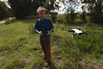 Man holding controller and flying quadcopter drone in field wearing orange helmet with face shield