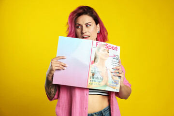 Beautiful woman with pink dyed hair holding sports magazine on yellow background