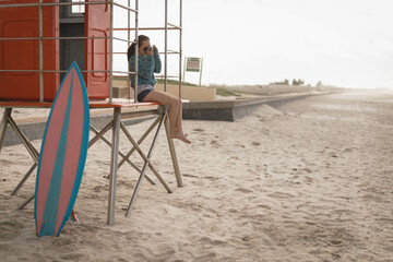 Fototapeta premium Red lifeguard stand is standing on sand near shore with pink-blue surfboard leaning