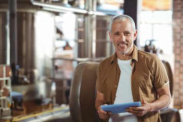 Senior male technician holding blue tablet and inspecting fermentation tanks and pipes at brewery