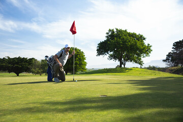 Male golfer kneeling on green holding flagstick, observing golf ball near hole, copy space