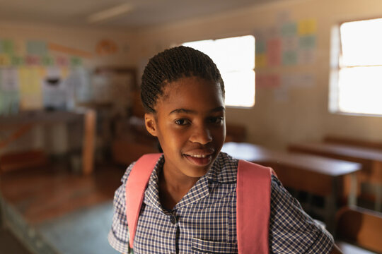 African American schoolgirl standing inside classroom wearing checkered uniform and pink backpack