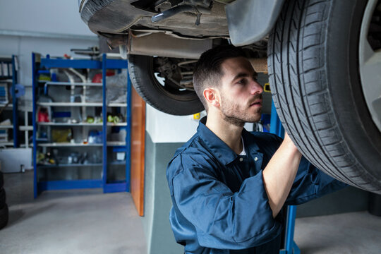 Male mechanic wearing coveralls inspecting car wheel under lift near workshop shelving, copy space