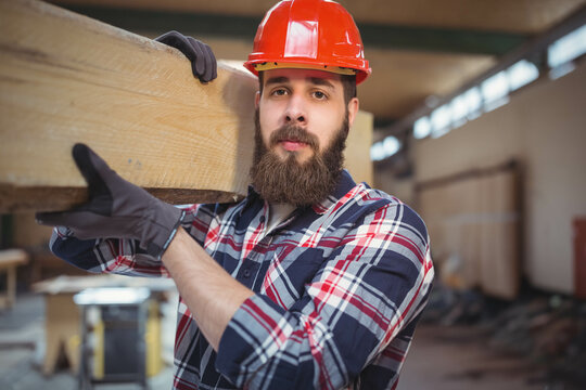 Long wooden beam resting on shoulder in woodworking shop with workbenches, tools and lumber stacks