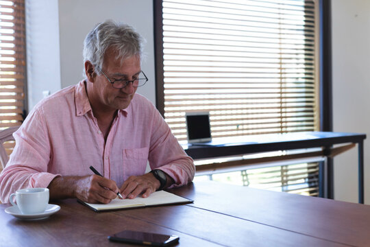 Senior man writing in open notebook at home workspace with pen coffee cup smartphone smartwatch - Powered by Adobe