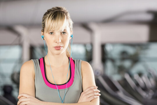 Woman standing in gym by treadmills wearing grey tank top, pink sports bra and blue earbuds