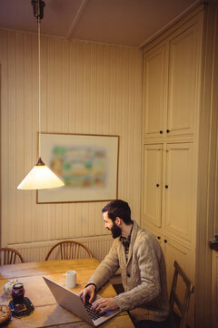 Bearded typing on laptop at wooden table with pendant lamp illuminating pastries and jars of jam