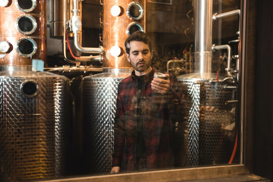 Man wearing shirt holding glass jar examining contents among copper stills and pipes in distillery