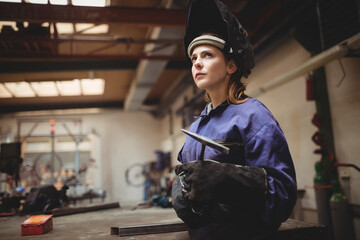 Female welder holding welding electrode at workbench wearing welding helmet and gloves, copy space