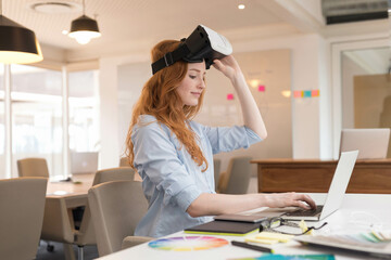 Red-haired woman sitting at office desk lifting virtual reality headset near laptop and color wheel