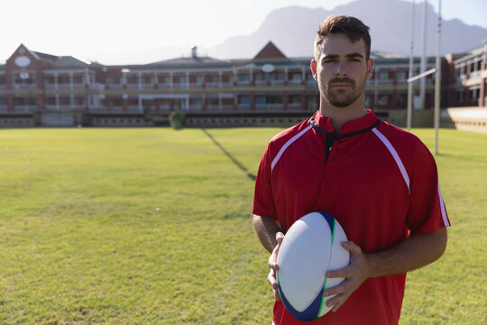 Male athlete standing on field in red jersey holding rugby ball near campus building, copy space