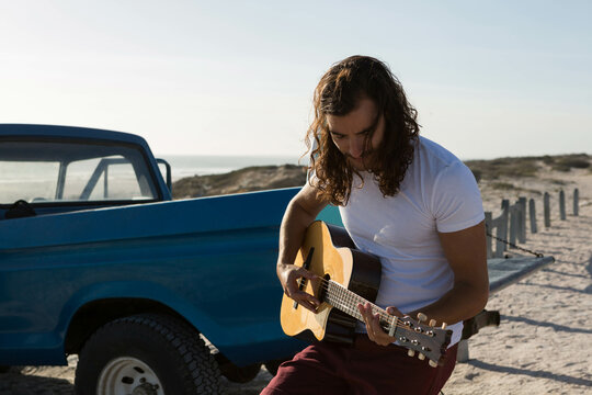 Guitarist strumming acoustic guitar on blue pickup truck by post and chain fence at sandy beach