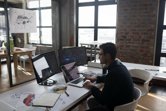 Laptop and dual monitors are displaying abstract car prototypes on white desk in design studio